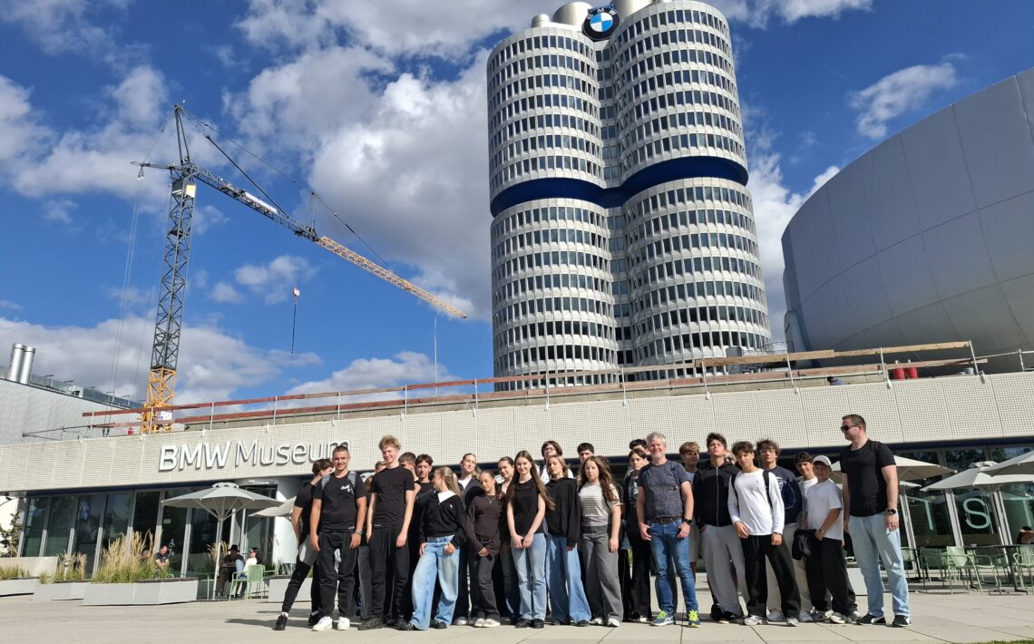 Grupa młodzieży i opiekunów pozuje przed BMW Museum w Monachium. W tle widać charakterystyczny wieżowiec BMW, logo marki oraz dźwig budowlany.