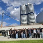 Grupa młodzieży i opiekunów pozuje przed BMW Museum w Monachium. W tle widać charakterystyczny wieżowiec BMW, logo marki oraz dźwig budowlany.