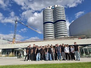 Grupa młodzieży i opiekunów pozuje przed BMW Museum w Monachium. W tle widać charakterystyczny wieżowiec BMW, logo marki oraz dźwig budowlany.