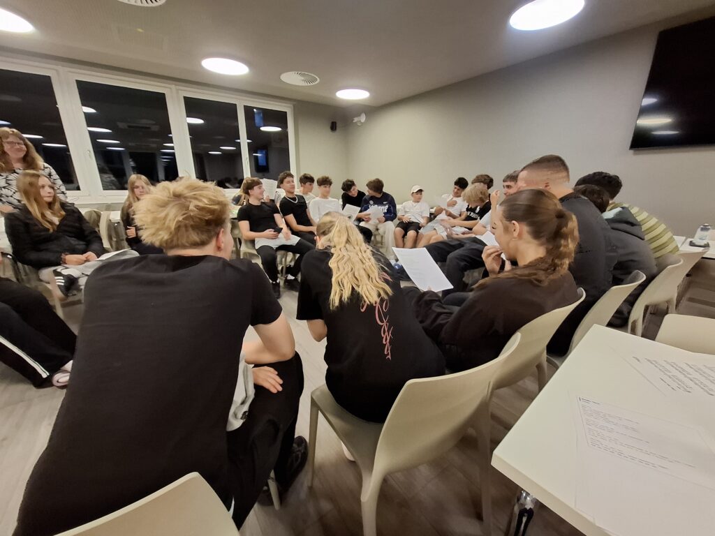 Grupa młodzieży i opiekunów siedzi w kręgu w jasnej sali. Uczestnicy trzymają kartki i wspólnie pracują lub rozmawiają.