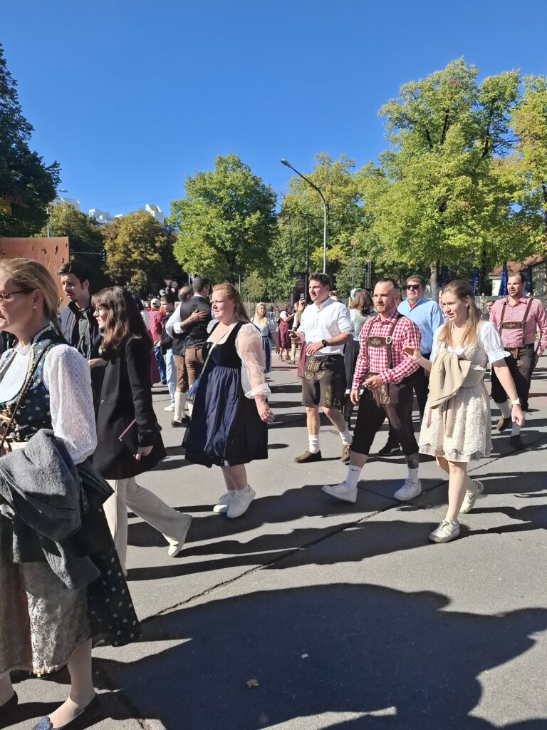 Uliczny pochód w słoneczny dzień. Wśród uczestników są osoby ubrane w tradycyjne bawarskie stroje, między innymi skórzane spodnie Lederhosen i sukienki w stylu Dirndl; w tle rosną zielone drzewa.