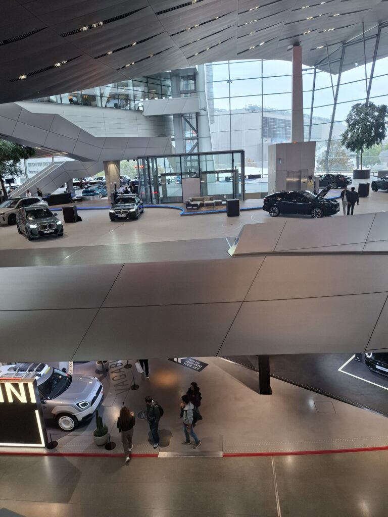 Wnętrze nowoczesnego obiektu BMW oglądane z wyższego poziomu. Na kilku kondygnacjach prezentowane są samochody, a przestrzeń wyróżniają przeszklone ściany i geometryczna architektura.