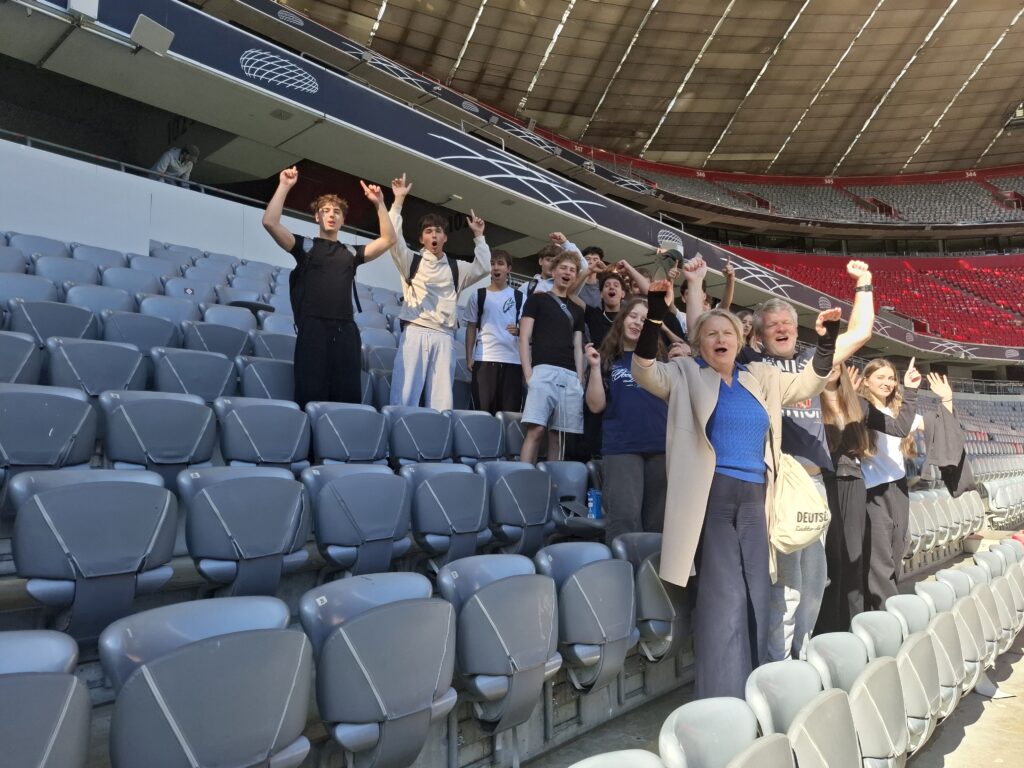 Młodzież wraz z opiekunami stoi na trybunach dużego stadionu. Uczestnicy uśmiechają się i unoszą ręce w geście radości; wokół znajdują się rzędy szarych i czerwonych siedzeń.