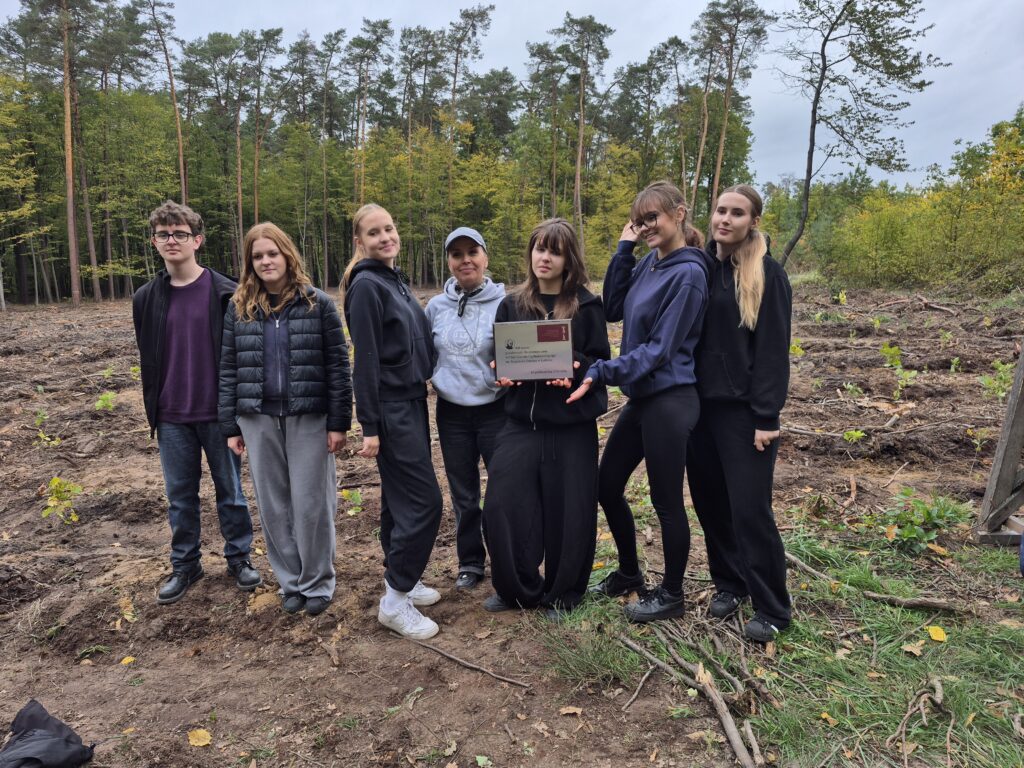 Sześcioro uczniów i dorosła opiekunka pozują na leśnej polanie. Dwie osoby trzymają pamiątkową tabliczkę z napisem dotyczącym posadzenia 440 drzew z okazji 440-lecia I Liceum Ogólnokształcącego im. Stanisława Staszica w Lublinie. W tle rosną młode drzewka.