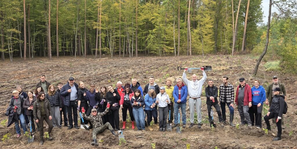 Duża grupa młodzieży i dorosłych pozuje na leśnej polanie po sadzeniu drzew. Uczestnicy uśmiechają się, machają rękami i trzymają łopaty. Jeden z mężczyzn unosi łopatę nad głową, a drugi siedzi z przodu z rozłożonymi ramionami. Wokół widoczne są rzędy młodych sadzonek.