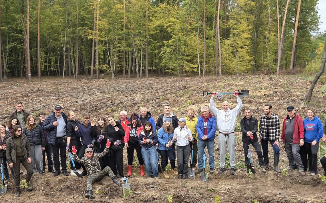 Duża grupa młodzieży i dorosłych pozuje na leśnej polanie po sadzeniu drzew. Uczestnicy uśmiechają się, machają rękami i trzymają łopaty. Jeden z mężczyzn unosi łopatę nad głową, a drugi siedzi z przodu z rozłożonymi ramionami. Wokół widoczne są rzędy młodych sadzonek.