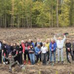 Duża grupa młodzieży i dorosłych pozuje na leśnej polanie po sadzeniu drzew. Uczestnicy uśmiechają się, machają rękami i trzymają łopaty. Jeden z mężczyzn unosi łopatę nad głową, a drugi siedzi z przodu z rozłożonymi ramionami. Wokół widoczne są rzędy młodych sadzonek.