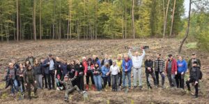 Duża grupa młodzieży i dorosłych pozuje na leśnej polanie po sadzeniu drzew. Uczestnicy uśmiechają się, machają rękami i trzymają łopaty. Jeden z mężczyzn unosi łopatę nad głową, a drugi siedzi z przodu z rozłożonymi ramionami. Wokół widoczne są rzędy młodych sadzonek.