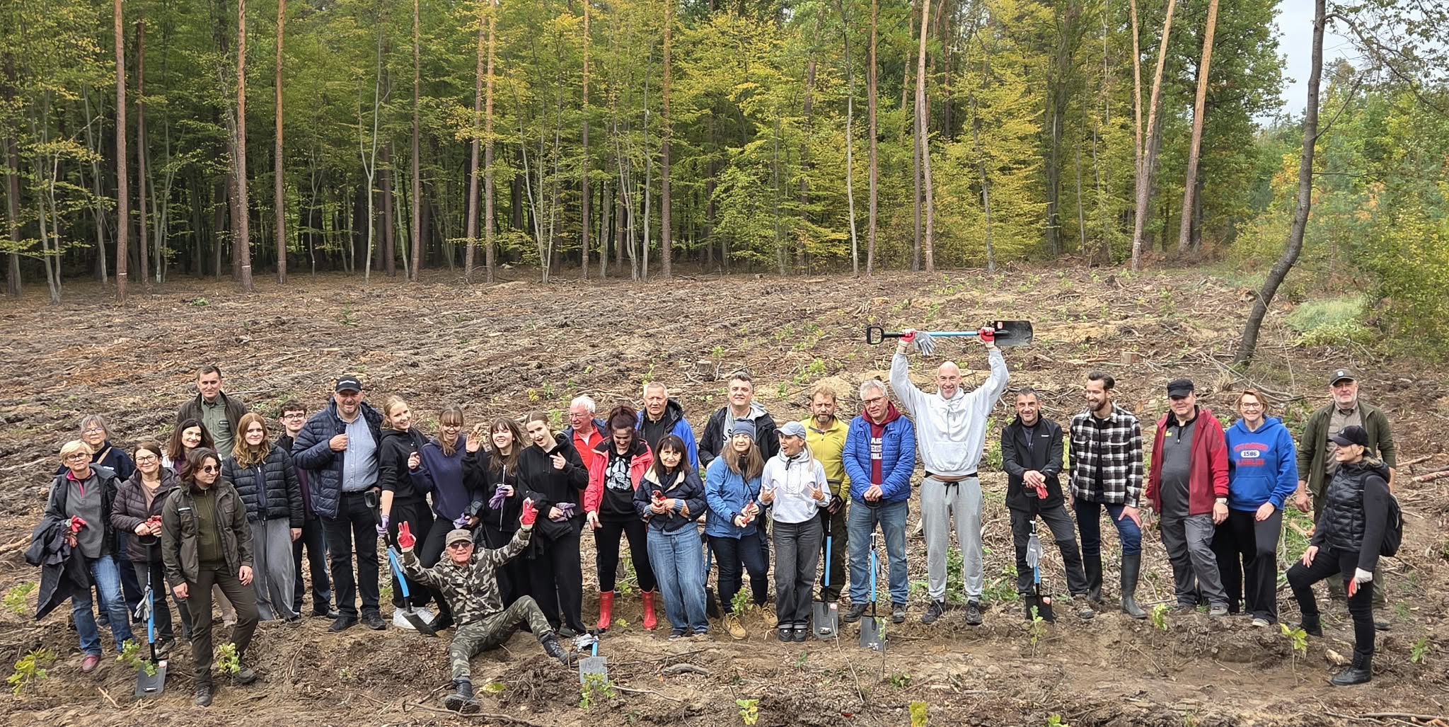 Duża grupa młodzieży i dorosłych pozuje na leśnej polanie po sadzeniu drzew. Uczestnicy uśmiechają się, machają rękami i trzymają łopaty. Jeden z mężczyzn unosi łopatę nad głową, a drugi siedzi z przodu z rozłożonymi ramionami. Wokół widoczne są rzędy młodych sadzonek.