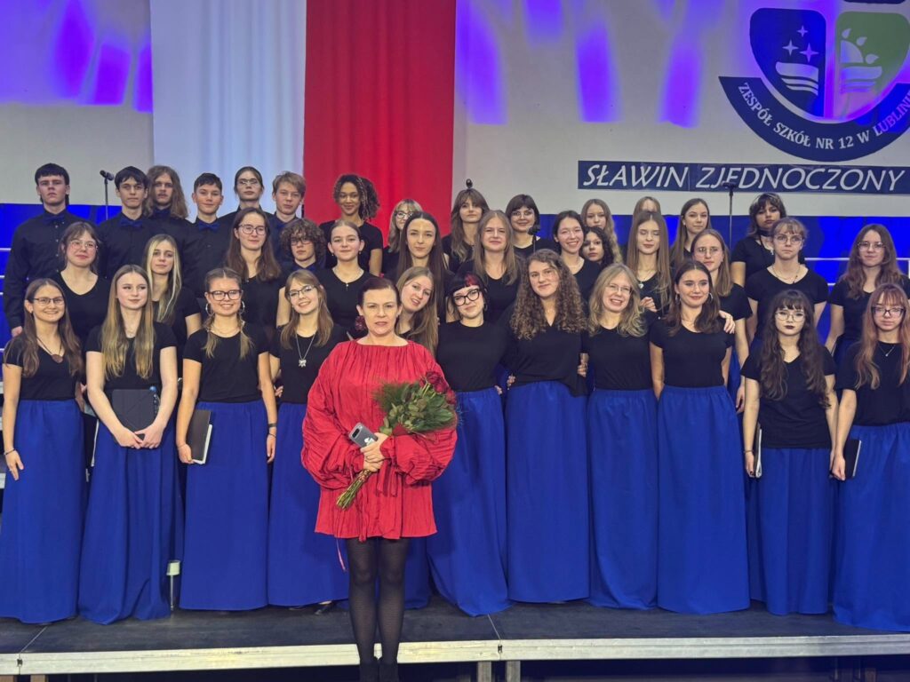 Zdjęcie grupowe chóru szkolnego na scenie. Młodzież ubrana jest w czarne koszulki i długie, niebieskie spódnice, a chłopcy w czarne koszule. Z przodu stoi kobieta w czerwonej sukience, trzymająca bukiet róż i telefon. W tle widoczne są biało-czerwone dekoracje, fioletowe oświetlenie, napis „SŁAWIN ZJEDNOCZONY” oraz fragment herbu Zespołu Szkół nr 12 w Lublinie.