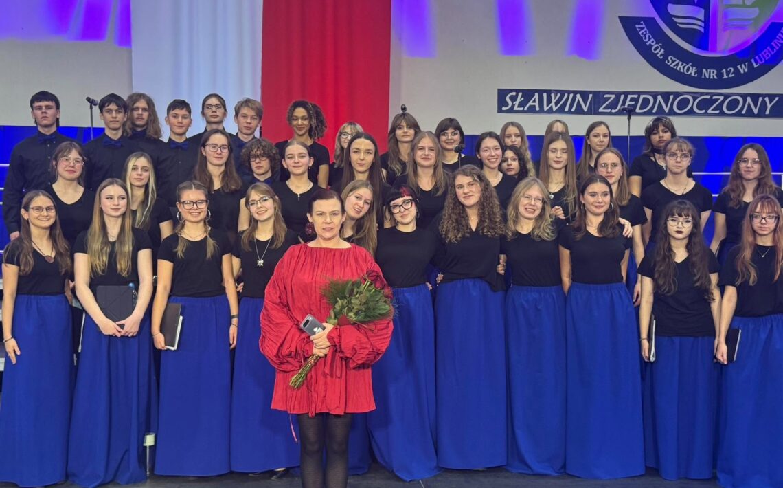 Zdjęcie grupowe chóru szkolnego na scenie. Młodzież ubrana jest w czarne koszulki i długie, niebieskie spódnice, a chłopcy w czarne koszule. Z przodu stoi kobieta w czerwonej sukience, trzymająca bukiet róż i telefon. W tle widoczne są biało-czerwone dekoracje, fioletowe oświetlenie, napis „SŁAWIN ZJEDNOCZONY” oraz fragment herbu Zespołu Szkół nr 12 w Lublinie.