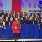 Zdjęcie grupowe chóru szkolnego na scenie. Młodzież ubrana jest w czarne koszulki i długie, niebieskie spódnice, a chłopcy w czarne koszule. Z przodu stoi kobieta w czerwonej sukience, trzymająca bukiet róż i telefon. W tle widoczne są biało-czerwone dekoracje, fioletowe oświetlenie, napis „SŁAWIN ZJEDNOCZONY” oraz fragment herbu Zespołu Szkół nr 12 w Lublinie.