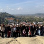 Duża grupa młodzieży pozuje do wspólnego zdjęcia na punkcie widokowym. W tle rozciąga się panorama miasteczka, lasów i szerokiej rzeki.