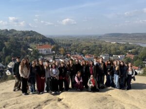 Duża grupa młodzieży pozuje do wspólnego zdjęcia na punkcie widokowym. W tle rozciąga się panorama miasteczka, lasów i szerokiej rzeki.