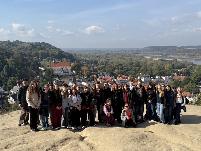 Duża grupa młodzieży pozuje do wspólnego zdjęcia na punkcie widokowym. W tle rozciąga się panorama miasteczka, lasów i szerokiej rzeki.