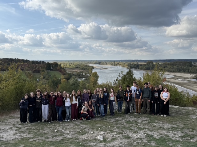 Grupa młodzieży pozuje na wysokim punkcie widokowym. Za nimi rozciąga się szeroka panorama rzeki, lasów i pól pod częściowo zachmurzonym niebem.