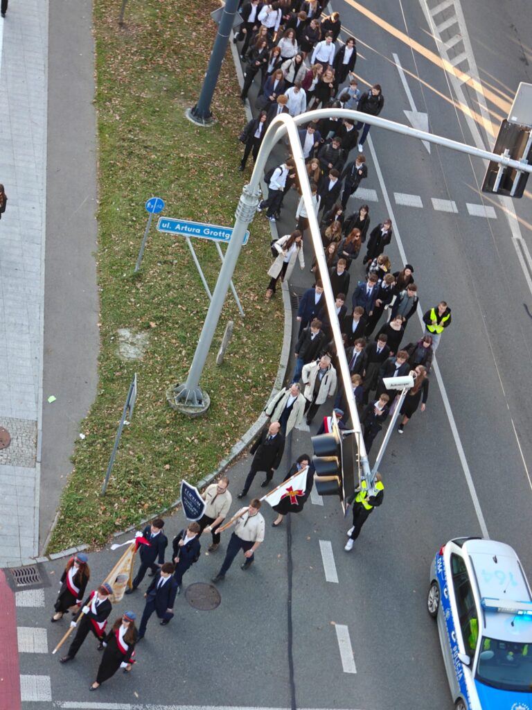 Widok z góry na uroczysty przemarsz uczniów I LO im. Stanisława Staszica w Lublinie. Na czele pochodu idzie poczet sztandarowy w ciemnych strojach z biało-czerwonymi szarfami. Za nim podążają uczniowie, nauczyciele i opiekunowie. Obok jedzie radiowóz policyjny.
