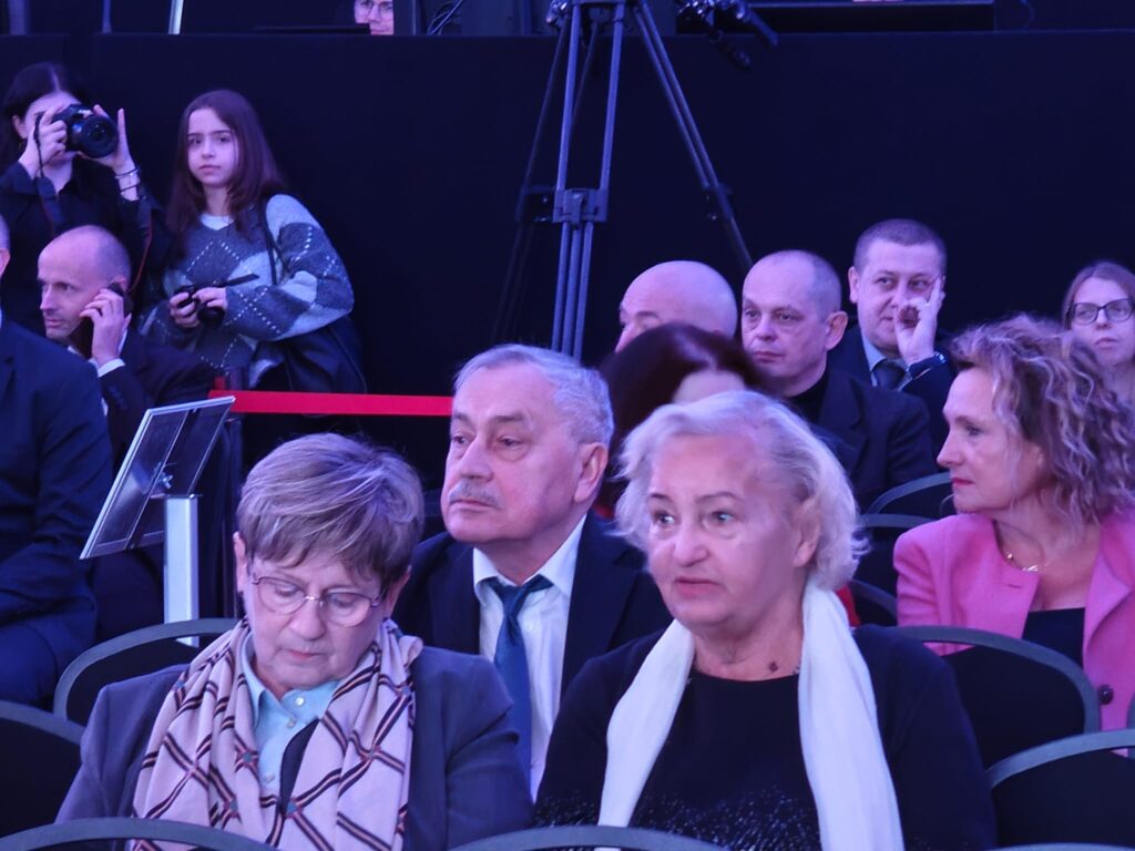 Zaproszeni goście siedzą w rzędach krzeseł podczas jubileuszu szkoły. Na pierwszym planie dwie starsze kobiety, jedna w okularach i wzorzystej apaszce, druga z białym szalem. Za nimi siedzą mężczyźni w garniturach. Salę oświetla niebiesko-fioletowe światło.