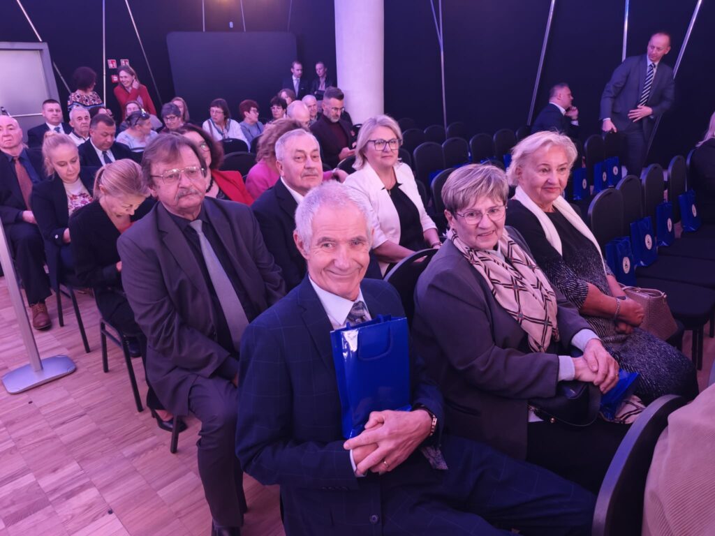 Grupa elegancko ubranych gości siedzących w sali widowiskowej. Na pierwszym planie uśmiechnięty siwowłosy mężczyzna w granatowym garniturze trzyma niebieską torbę z upominkiem. Obok siedzą kobiety w wizytowych strojach. W tle widoczne są kolejne rzędy uczestników.