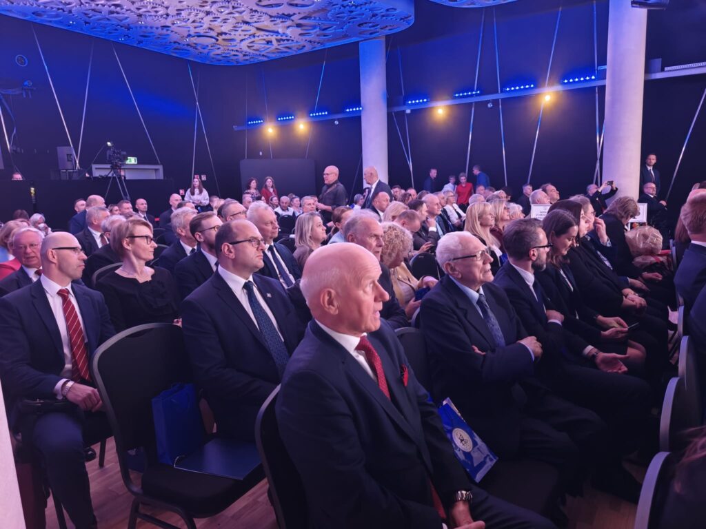 Zaproszeni goście jubileuszu 440-lecia I LO im. Stanisława Staszica w Lublinie siedzą w rzędach krzeseł w sali widowiskowej. Większość osób ubrana jest w eleganckie garnitury i stroje wizytowe. Na pierwszym planie widoczni są mężczyźni w ciemnych garniturach, a w tle liczni uczestnicy uroczystości. Salę oświetla niebiesko-fioletowe światło.
