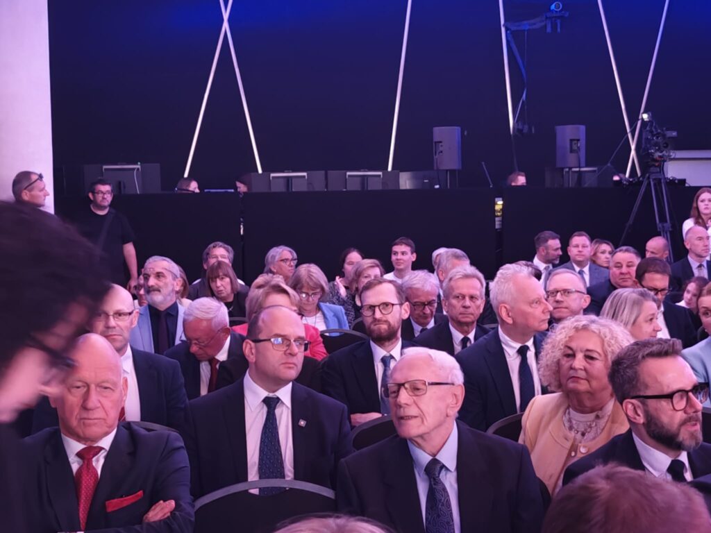 Uczestnicy jubileuszu 440-lecia I LO im. Stanisława Staszica w Lublinie siedzą w sali widowiskowej. Na pierwszym planie mężczyźni w eleganckich garniturach, za nimi kolejne rzędy gości. Wnętrze oświetlone jest fioletowo-niebieskim światłem.