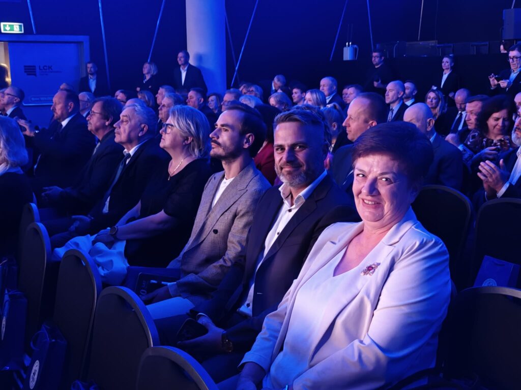 Goście jubileuszu siedzą w rzędach krzeseł, obserwując uroczystość. Na pierwszym planie uśmiechnięta kobieta w jasnym żakiecie oraz mężczyźni w eleganckich marynarkach. Salę wypełnia intensywne niebieskie oświetlenie.