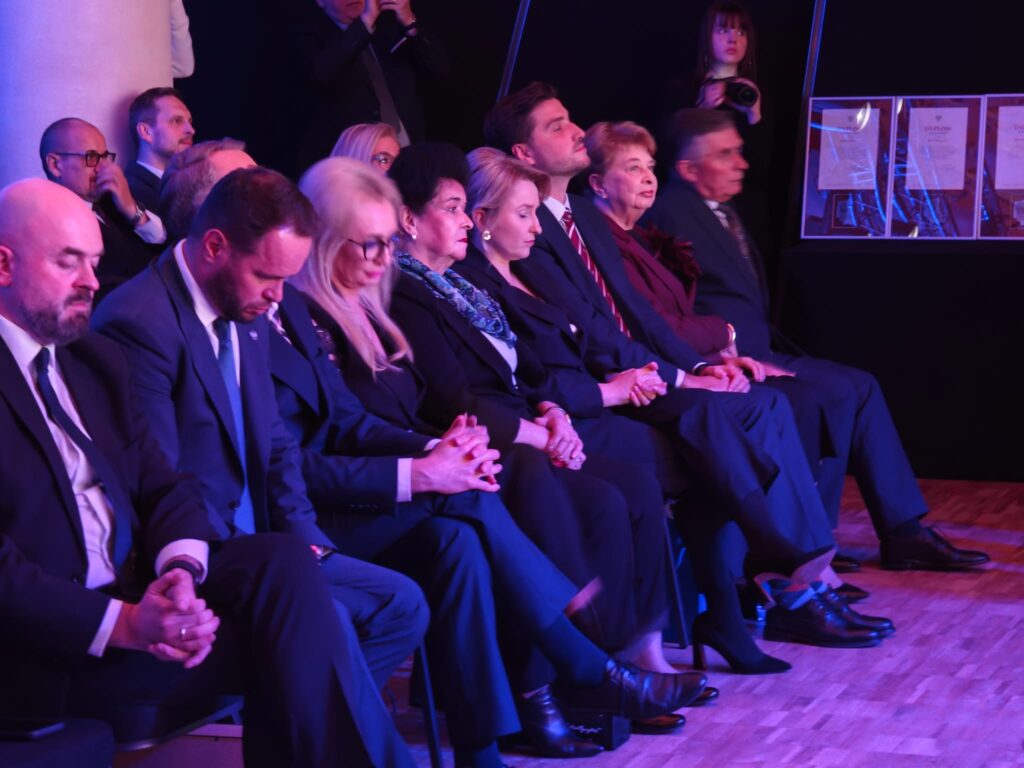 Elegancko ubrani uczestnicy jubileuszu siedzą w pierwszym rzędzie, zwróceni w stronę sceny. Wśród nich kobiety w ciemnych strojach oraz mężczyźni w garniturach. W tle widoczne są oprawione dyplomy.