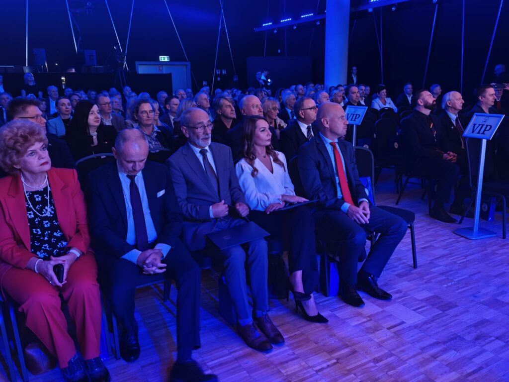 Widok na publiczność podczas uroczystości jubileuszowej. W pierwszym rzędzie siedzą kobieta w czerwonym kostiumie oraz elegancko ubrani goście. Po prawej stronie widoczne są tabliczki z napisem „VIP”.