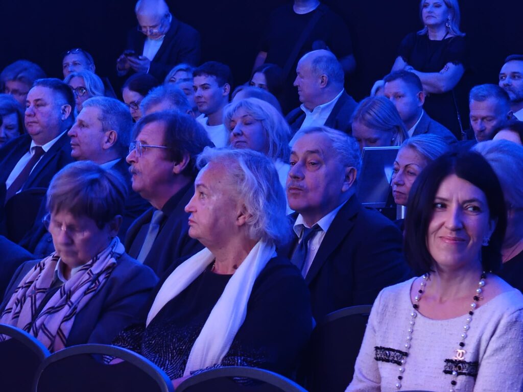 Zbliżenie na gości siedzących w kilku rzędach. Na pierwszym planie starsze kobiety i mężczyźni w eleganckich strojach oraz uśmiechnięta kobieta w jasnej bluzce z naszyjnikiem. Całą salę oświetla intensywne niebieskie światło.