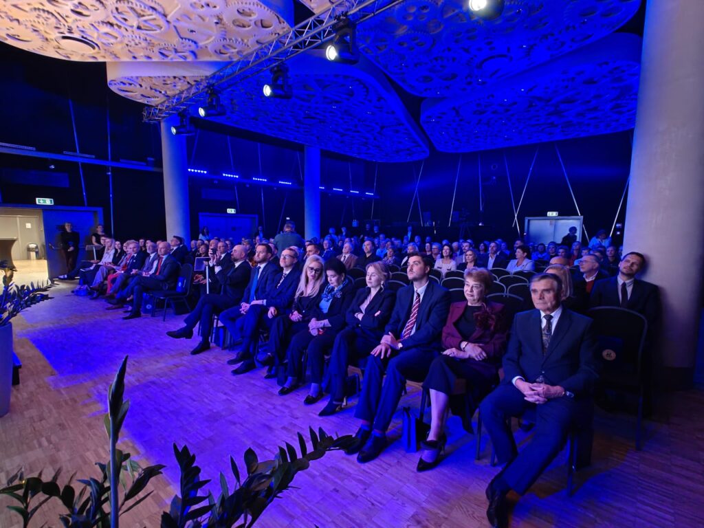 Goście jubileuszu 440-lecia I LO im. Stanisława Staszica w Lublinie siedzą w rzędach krzeseł, obserwując uroczystość. Sala jest oświetlona niebieskim światłem, a sufit zdobią dekoracyjne wzory przypominające koła zębate.