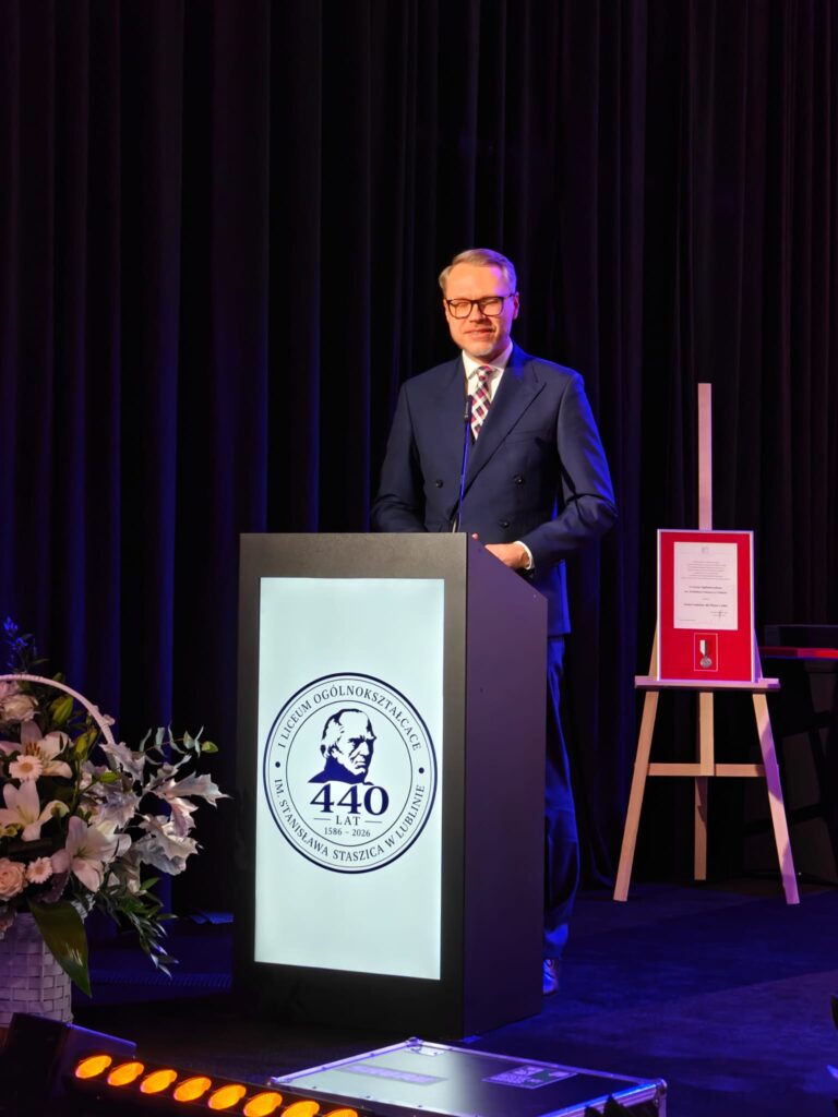 Mężczyzna z krótką brodą, ubrany w granatowy garnitur i niebieski krawat, przemawia przy mównicy z logo jubileuszu. Podczas wystąpienia gestykuluje dłonią.