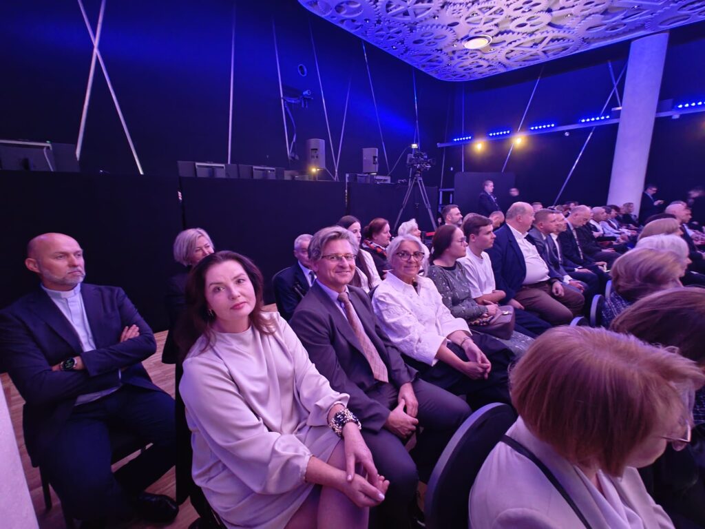 Uczestnicy jubileuszu siedzą w sali oświetlonej niebieskim światłem. Na pierwszym planie kobieta w jasnej sukience, mężczyzna w szarym garniturze oraz starsza kobieta w białej bluzce. Wśród gości widoczny jest również duchowny w koloratce.