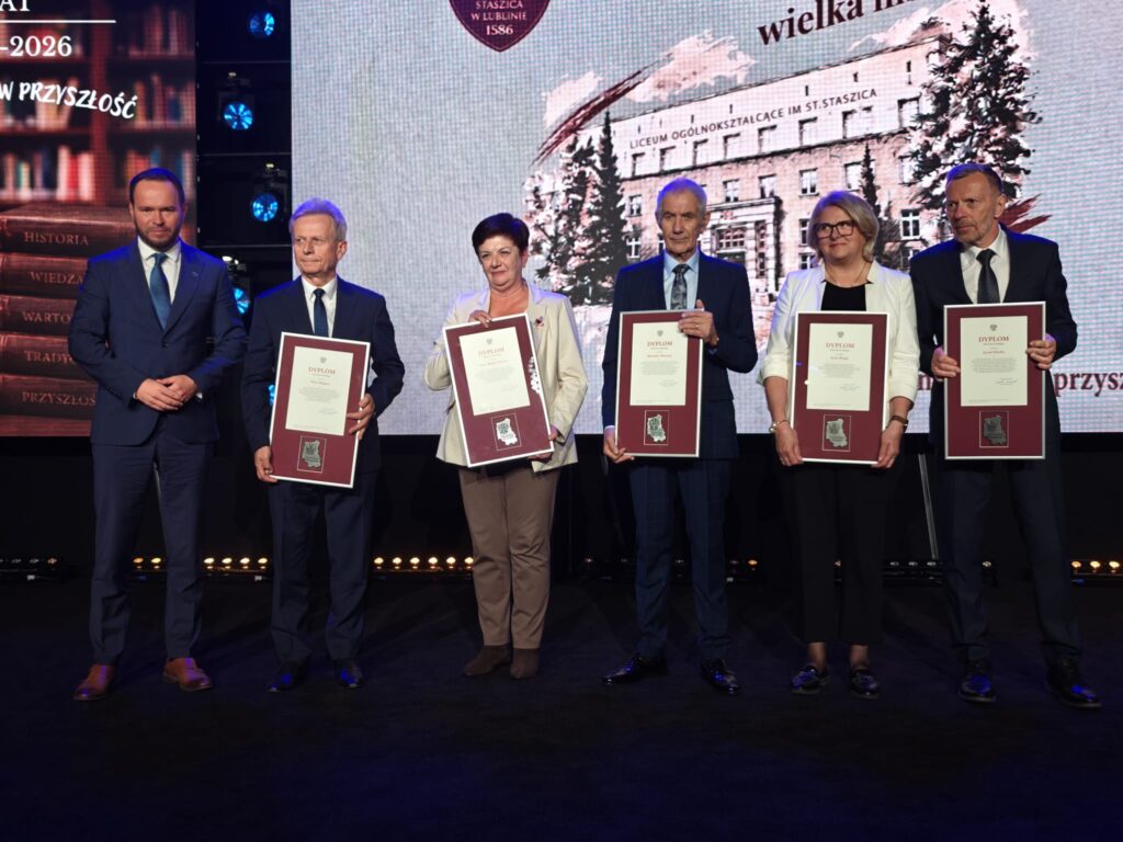 Sześć osób stoi na scenie podczas jubileuszu 440-lecia I LO im. Stanisława Staszica w Lublinie. Pięcioro uczestników prezentuje oprawione dyplomy w bordowych ramach. W tle widoczna jest grafika przedstawiająca budynek szkoły.