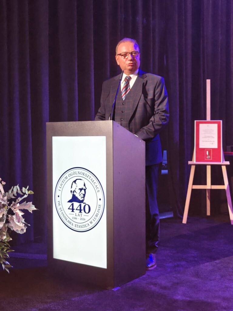 Mężczyzna w okularach, ciemnym garniturze w kratę i pasiastym krawacie przemawia przy mównicy z jubileuszowym logo szkoły. Obok znajduje się oprawiony dokument z medalem.