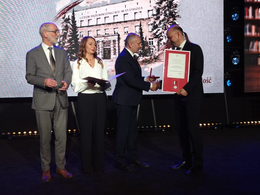 Na scenie dwóch mężczyzn w ciemnych garniturach podaje sobie dłonie. Jeden trzyma oprawiony dyplom z medalem. Obok stoją kobieta w białej bluzce z otwartą teczką oraz starszy mężczyzna w szarym garniturze.