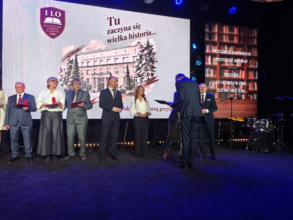 Grupa elegancko ubranych osób uczestniczy w ceremonii wręczania wyróżnień na scenie. Część trzyma czerwone etui i dokumenty. W tle wyświetlono budynek szkoły oraz hasło „Tu zaczyna się wielka historia…”.