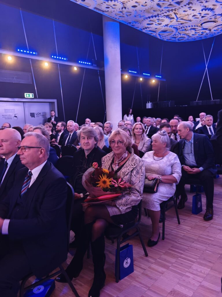 Elegancko ubrani goście siedzą w rzędach krzeseł w sali o niebieskim oświetleniu. Na pierwszym planie kobieta w okularach trzyma bukiet kwiatów ze słonecznikiem. Wśród publiczności widoczne są osoby rozmawiające i obserwujące wydarzenie.