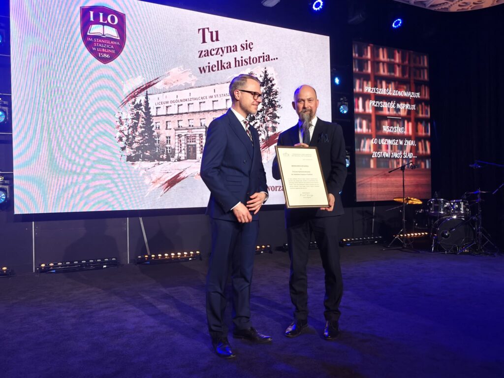 Dwóch mężczyzn w granatowych garniturach stoi na scenie. Mężczyzna z długą siwą brodą prezentuje oprawiony dokument z napisem „Dyplom uznania”. W tle znajduje się grafika budynku szkoły oraz jubileuszowe hasło „Tu zaczyna się wielka historia…”.