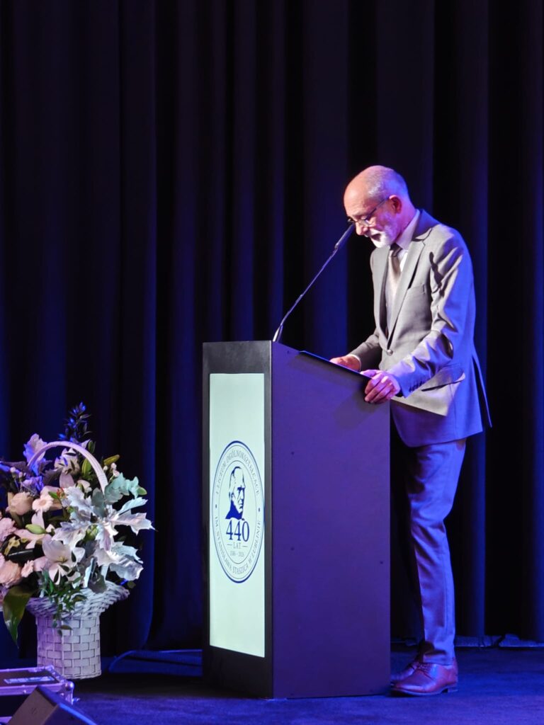 Starszy, łysy mężczyzna z siwą brodą i okularami, ubrany w jasnoszary garnitur, przemawia przy mównicy z logo 440-lecia I LO im. Stanisława Staszica w Lublinie. Obok stoi kosz z białymi kwiatami.