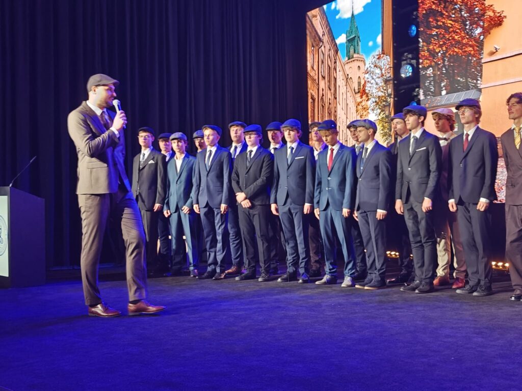 Na scenie stoi grupa kilkunastu młodych mężczyzn w eleganckich garniturach i kaszkietach. Po lewej mężczyzna w szarym garniturze mówi do mikrofonu. W tle wyświetlono fotografie zabytkowych budynków Lublina.