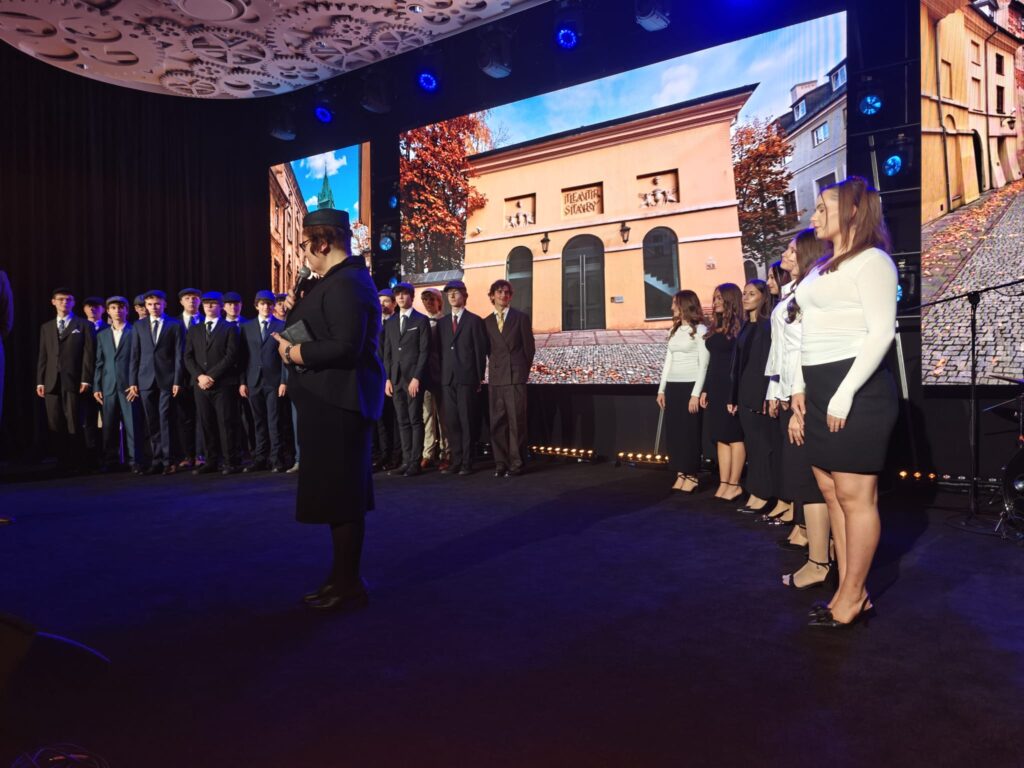 Na scenie stoją dwie grupy młodzieży: po lewej młodzi mężczyźni w garniturach i kaszkietach, po prawej młode kobiety w biało-czarnych strojach. Na środku kobieta w czarnym ubraniu przemawia do mikrofonu. W tle wyświetlono fotografię budynku Teatru Starego w Lublinie.