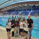 Na zdjęciu grupa siedmiu młodych zawodniczek i trener pozują przy basenie sportowym. Część dziewcząt ma na szyjach medale, jedna trzyma dyplom, a druga unosi puchar. W tle widać turkusową wodę basenu, tory pływackie oraz trybuny z niebieskimi siedzeniami.