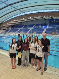 Na zdjęciu grupa siedmiu młodych zawodniczek i trener pozują przy basenie sportowym. Część dziewcząt ma na szyjach medale, jedna trzyma dyplom, a druga unosi puchar. W tle widać turkusową wodę basenu, tory pływackie oraz trybuny z niebieskimi siedzeniami.