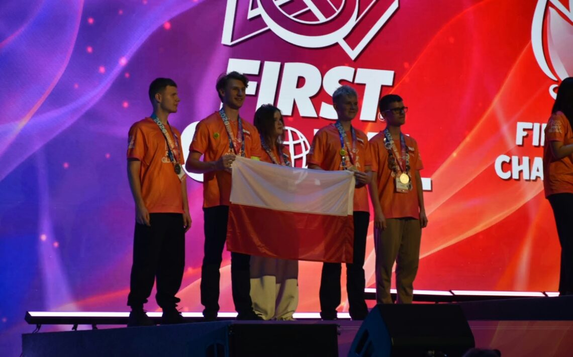 Pięcioro reprezentantów Polski pozuje na scenie podczas ceremonii zawodów FIRST Global. Uczestnicy mają pomarańczowe koszulki i medale zawieszone na szyjach. Dwie osoby trzymają biało-czerwoną flagę Polski. Za nimi znajduje się wielki ekran z logo wydarzenia na czerwono-fioletowym tle.
