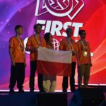 Pięcioro reprezentantów Polski pozuje na scenie podczas ceremonii zawodów FIRST Global. Uczestnicy mają pomarańczowe koszulki i medale zawieszone na szyjach. Dwie osoby trzymają biało-czerwoną flagę Polski. Za nimi znajduje się wielki ekran z logo wydarzenia na czerwono-fioletowym tle.