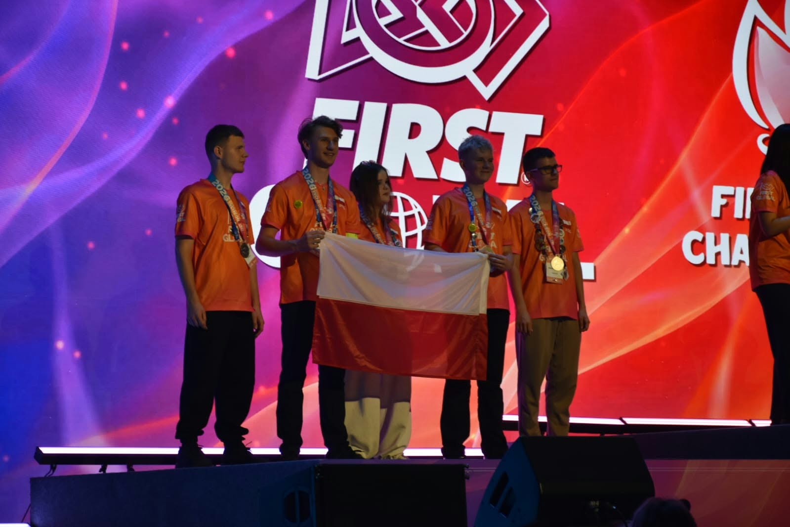Pięcioro reprezentantów Polski pozuje na scenie podczas ceremonii zawodów FIRST Global. Uczestnicy mają pomarańczowe koszulki i medale zawieszone na szyjach. Dwie osoby trzymają biało-czerwoną flagę Polski. Za nimi znajduje się wielki ekran z logo wydarzenia na czerwono-fioletowym tle.