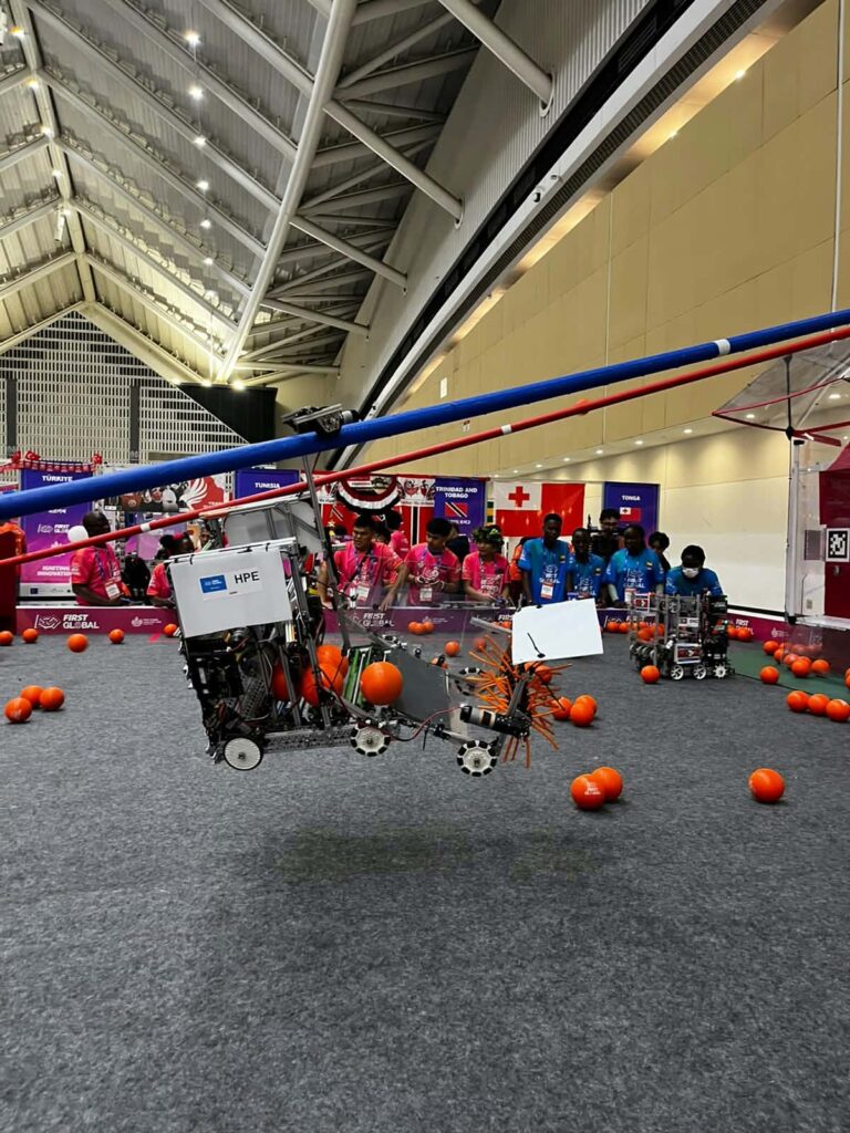 Robot z oznaczeniem „HPE”, wyposażony w metalowe ramiona i mechanizm z pomarańczowymi elementami, znajduje się częściowo nad podłożem areny. Wokół leżą pomarańczowe piłki. W tle uczestnicy obserwują przebieg konkurencji.