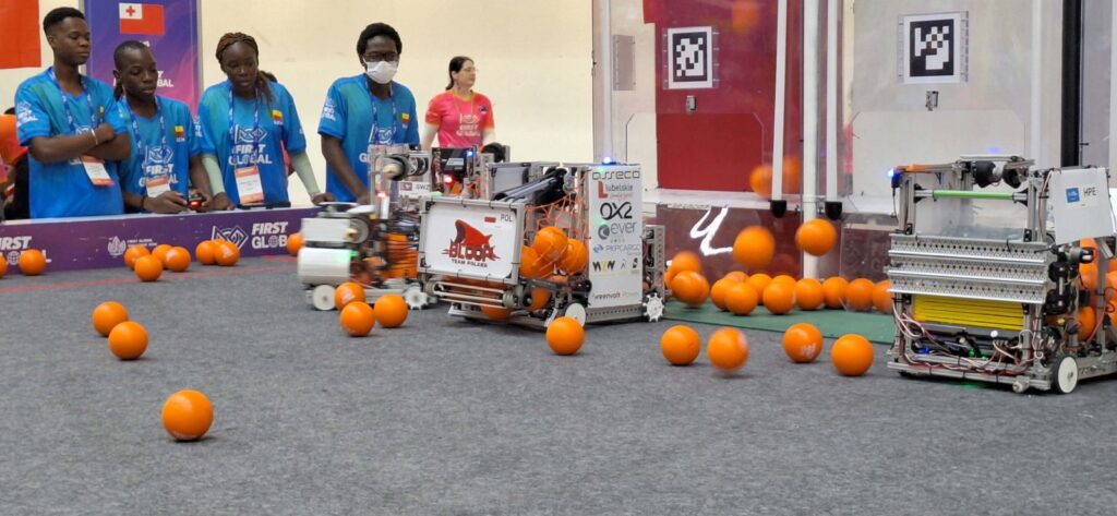 Na szarej arenie rywalizują roboty z metalowymi konstrukcjami. Wśród licznych pomarańczowych piłek znajduje się polski robot z oznaczeniem „POL” i logo „BŁOCK Team Poland”. Za barierą stoją uczestnicy w niebieskich koszulkach.