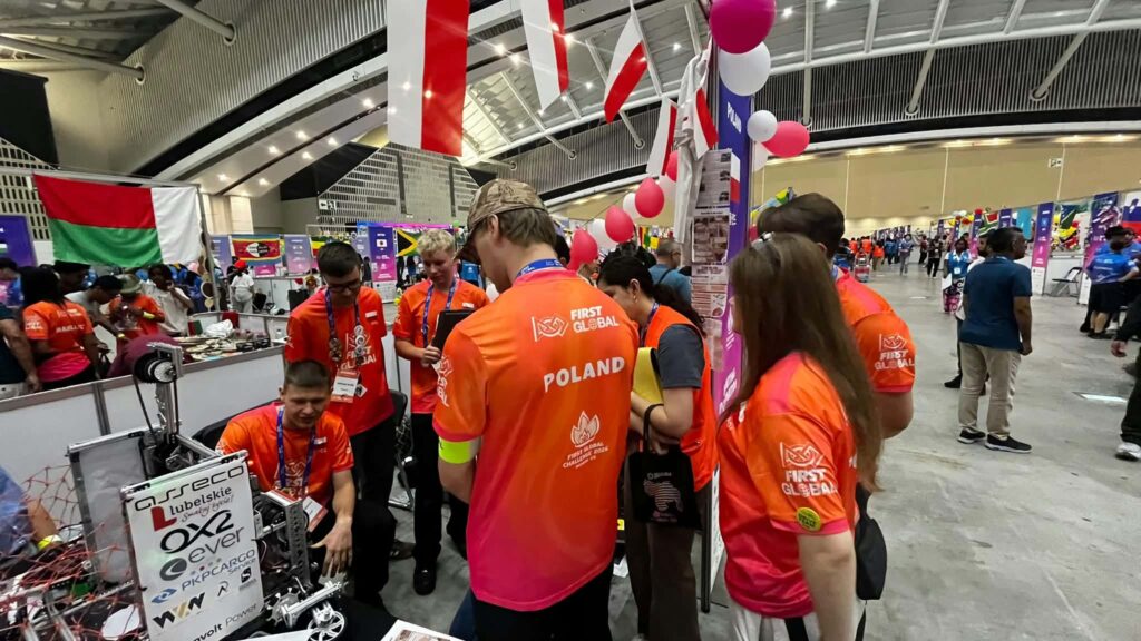 Uczestnicy polskiej drużyny w pomarańczowych koszulkach z napisami „FIRST GLOBAL” i „POLAND” pracują przy robocie konkursowym. W tle widoczne są stanowiska innych państw, flagi narodowe oraz biało-czerwone balony.