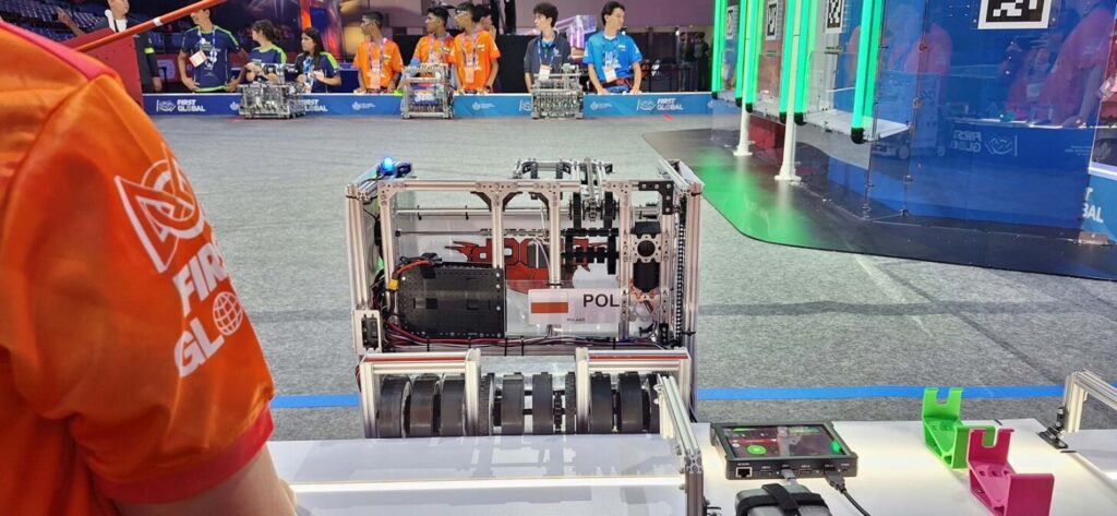 Na pierwszym planie stoi robot z metalową ramą, kołami i oznaczeniem „POL” oraz polską flagą. Obok znajduje się panel sterujący. W tle uczestnicy różnych drużyn obserwują arenę zawodów FIRST Global.
